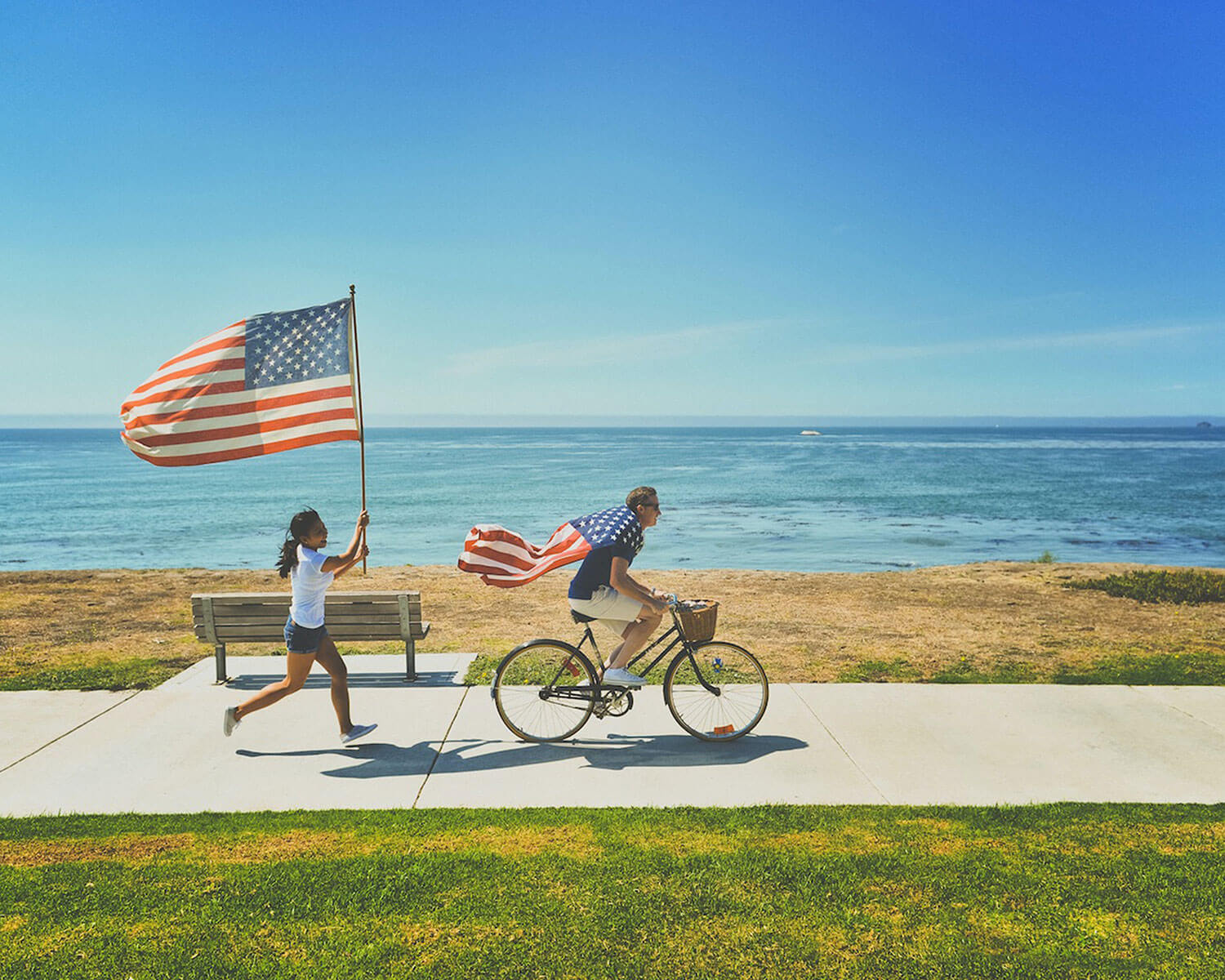 A man riding a bike wearing the American flag like a cape around his shoulders followed by a woman waving a large American flag running behind him