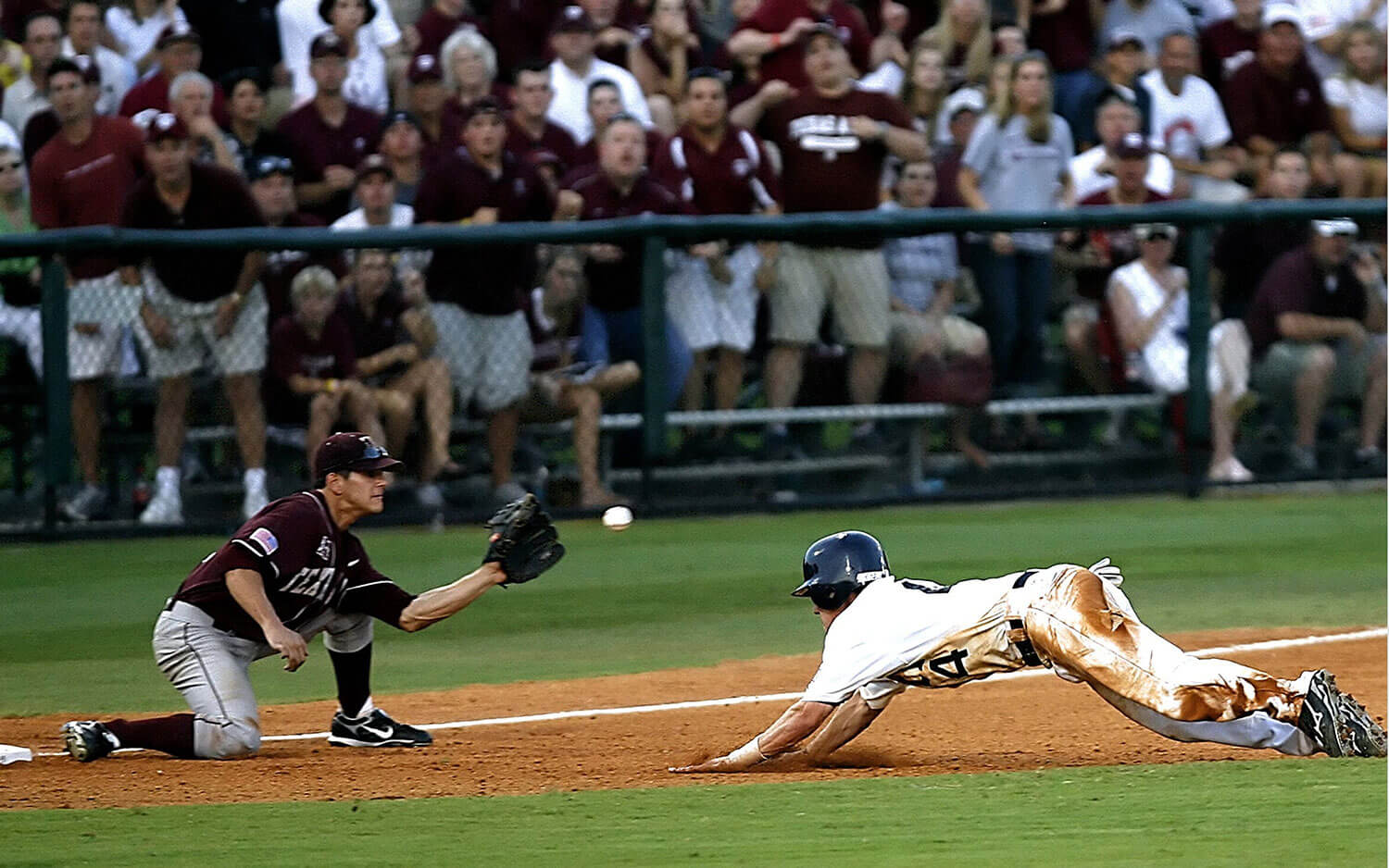 Baseball player sliding headfirst to make it to base with ball mid air about to land in the baseman's glove. Crowd cheering in the background