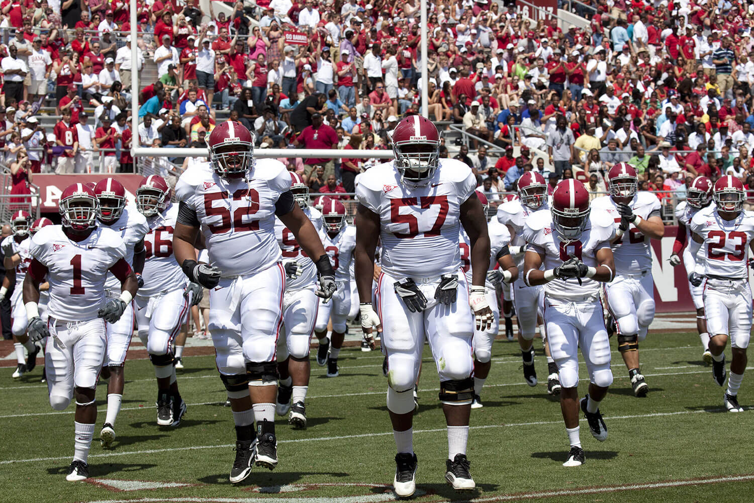 American Football team on the field dressed in white with Maroon numbers. Full stadium of fans also dressed in Maroon behind