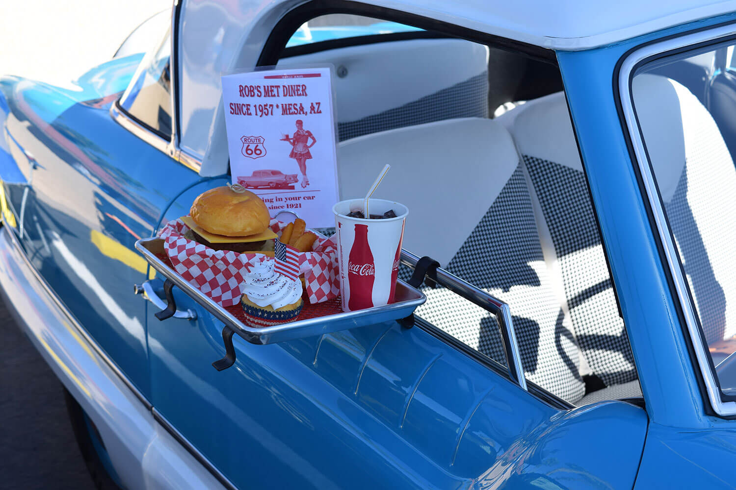 A blue car from the 50s parked at an American diner with an American style meal of hamburger, fries and a coke set on a tray hanging from the open window